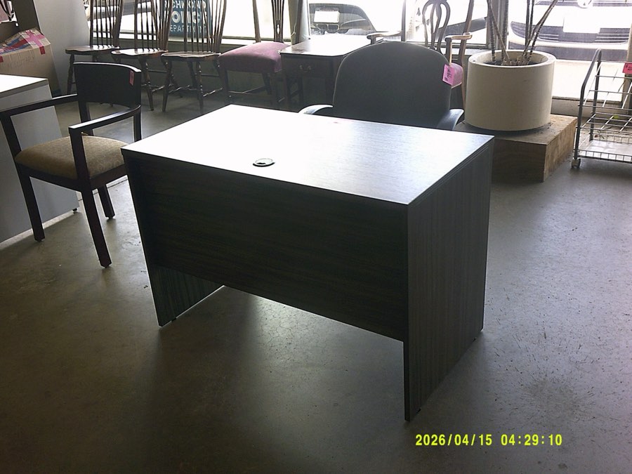 A compact dark laminate desk shell with a grommeted top, photographed near the store windows.