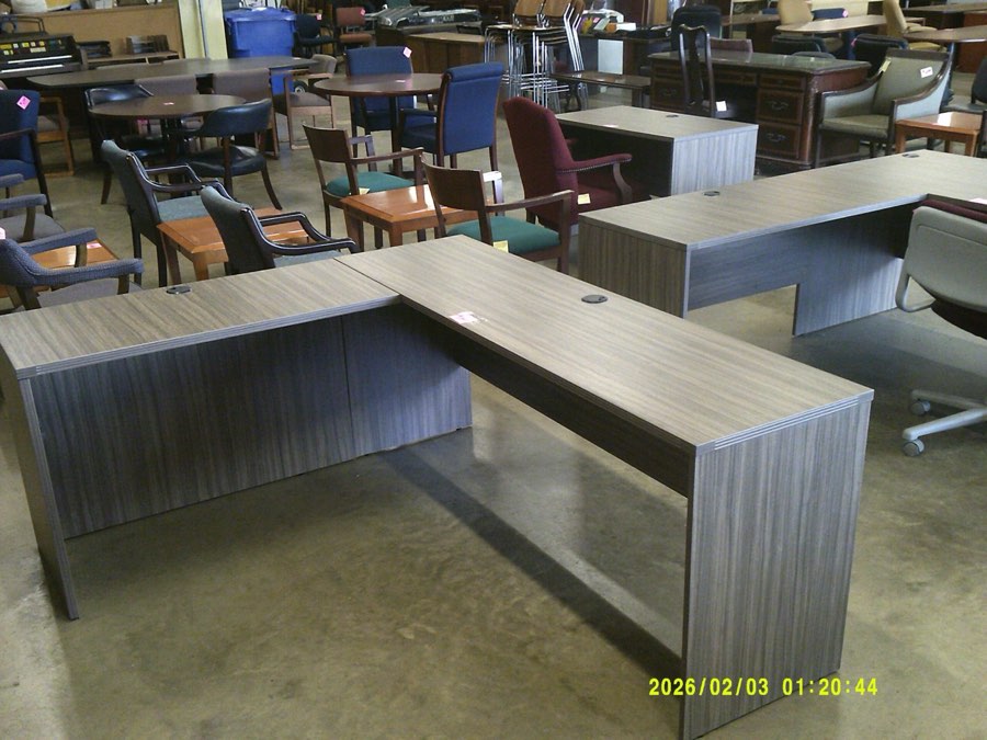 Two grey laminate L-shaped desk shells on the office furniture floor, with used chairs and tables behind them.